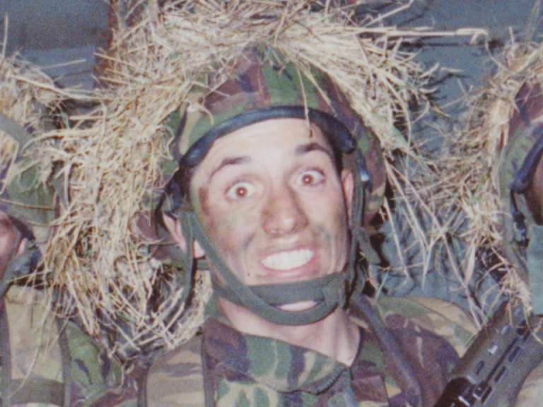 Grinning soldier in camouflage gear with foliage headgear, posing for the camera.