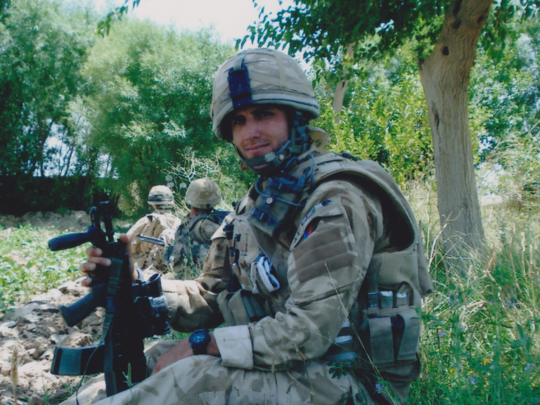 Soldier in military uniform with a rifle, sitting among trees and fellow soldiers.