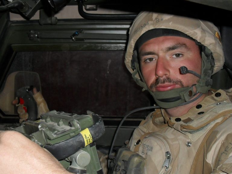 Soldier in a military vehicle wearing a helmet and tactical gear.