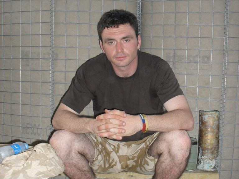 Man sitting on the ground, wearing a dark t-shirt and shorts, with hands clasped.