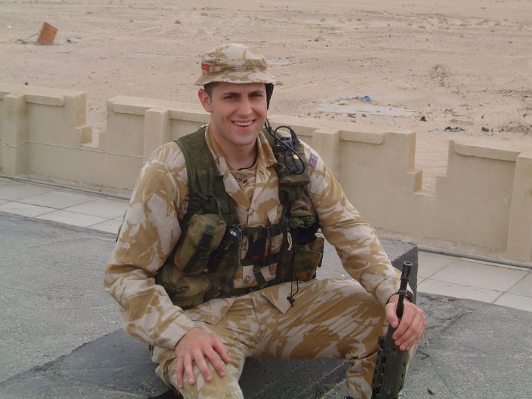 A soldier in camouflage uniform sitting on a surface, smiling in a sandy environment.