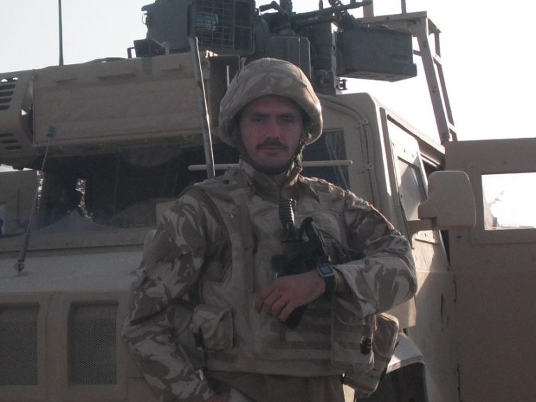 Soldier in military uniform posing in front of an armoured vehicle.